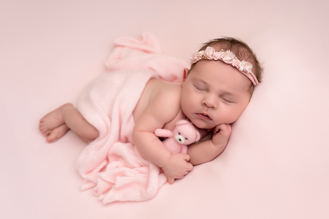 newborn girl on pink background in side lay pose wearing delicate pink flower headband and holding needle felted teddy bear